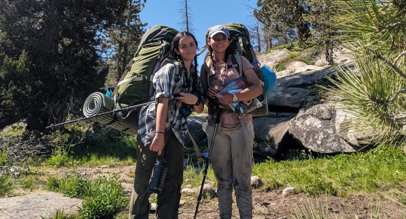 Two people wearing backpacks stand amongst trees and boulders and smile for the photo. 